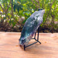 A polished natural crystal slab with a blue-green and purple marbled pattern, displayed on a small black metal stand on a wooden surface with a blurred green garden background.