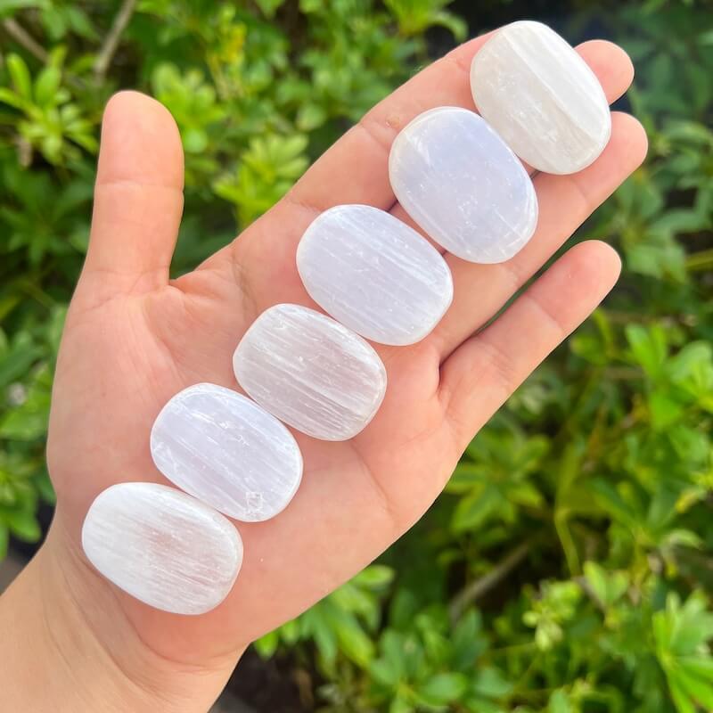 Five smooth, polished white Selenite gemstone pendants arranged on an open hand, with a blurred green garden background.