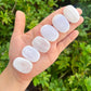 Five polished white selenite stones arranged on an open hand, with a blurred green garden background.