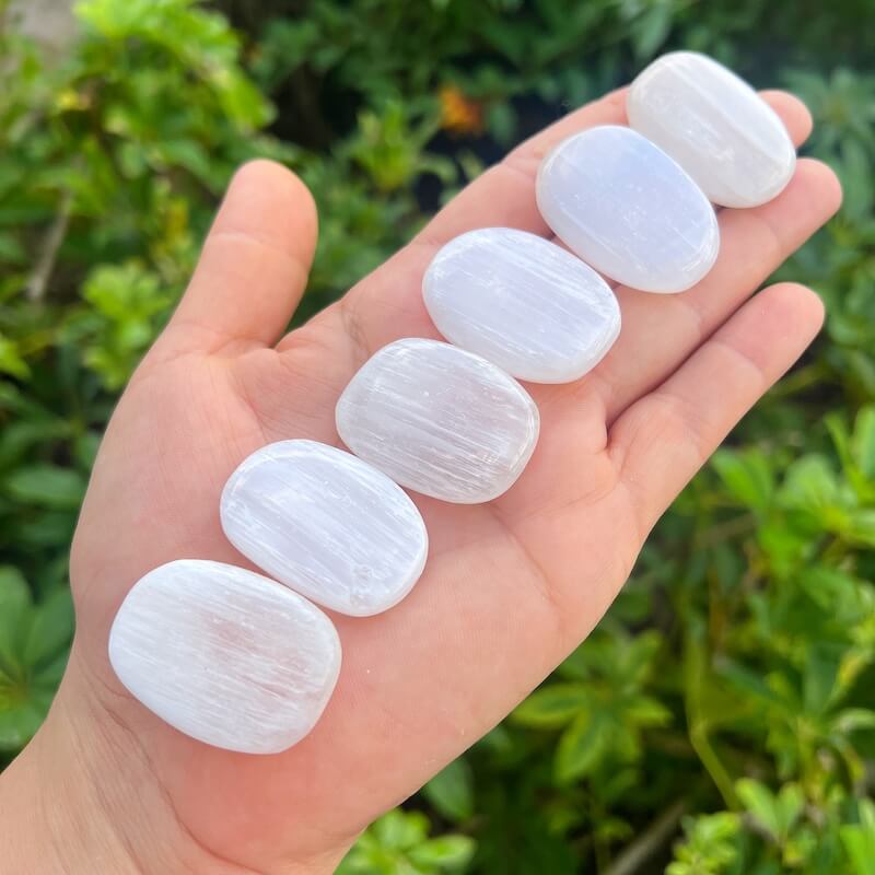 Five polished white stone pendants with a subtle pearlescent sheen are held in an open hand, with a blurred background of green foliage.